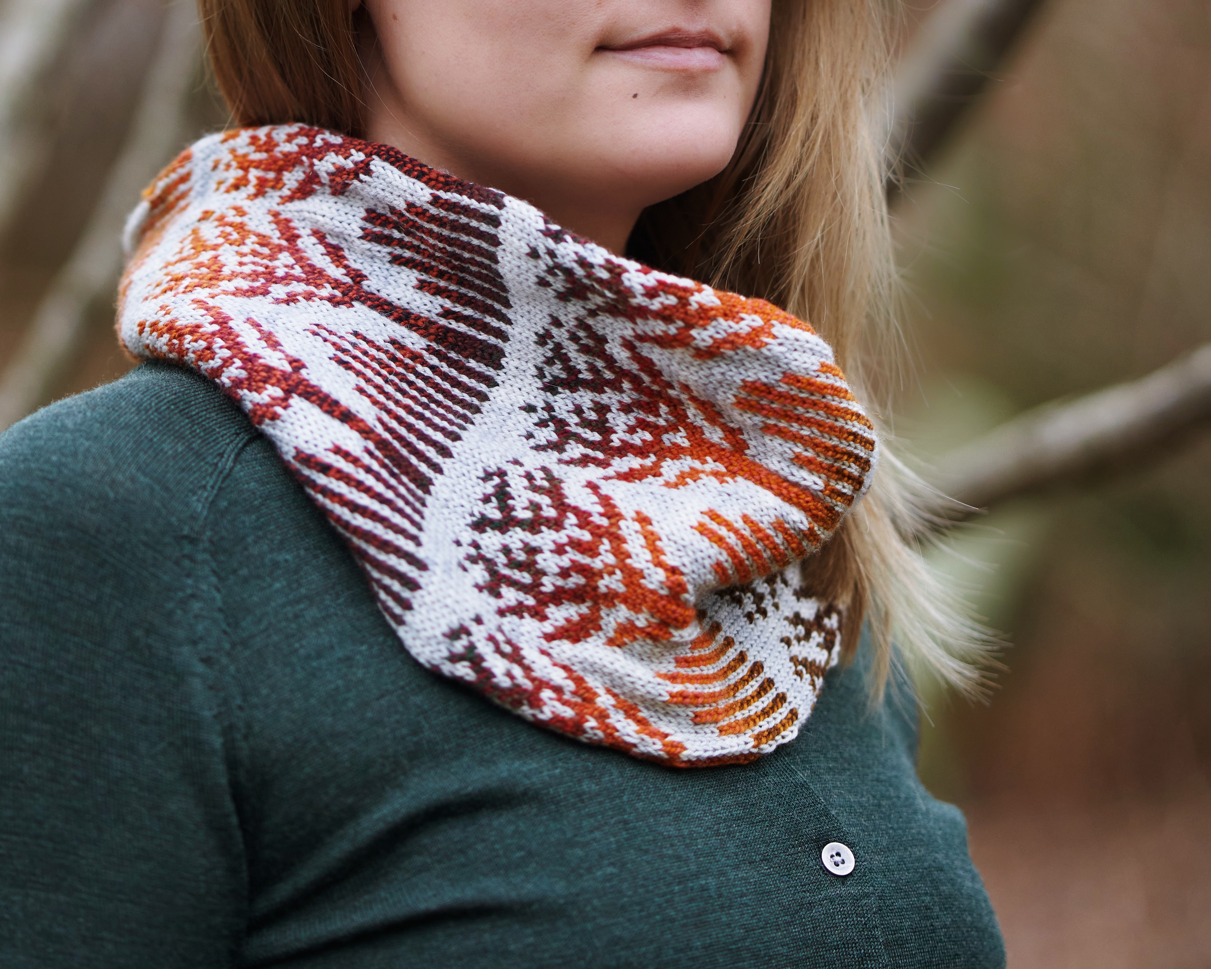 Closeup of a hank nit cowl worn on the neck of a blonde woman. The cowl features intricate colorwork in fall colors with a pale gray background.