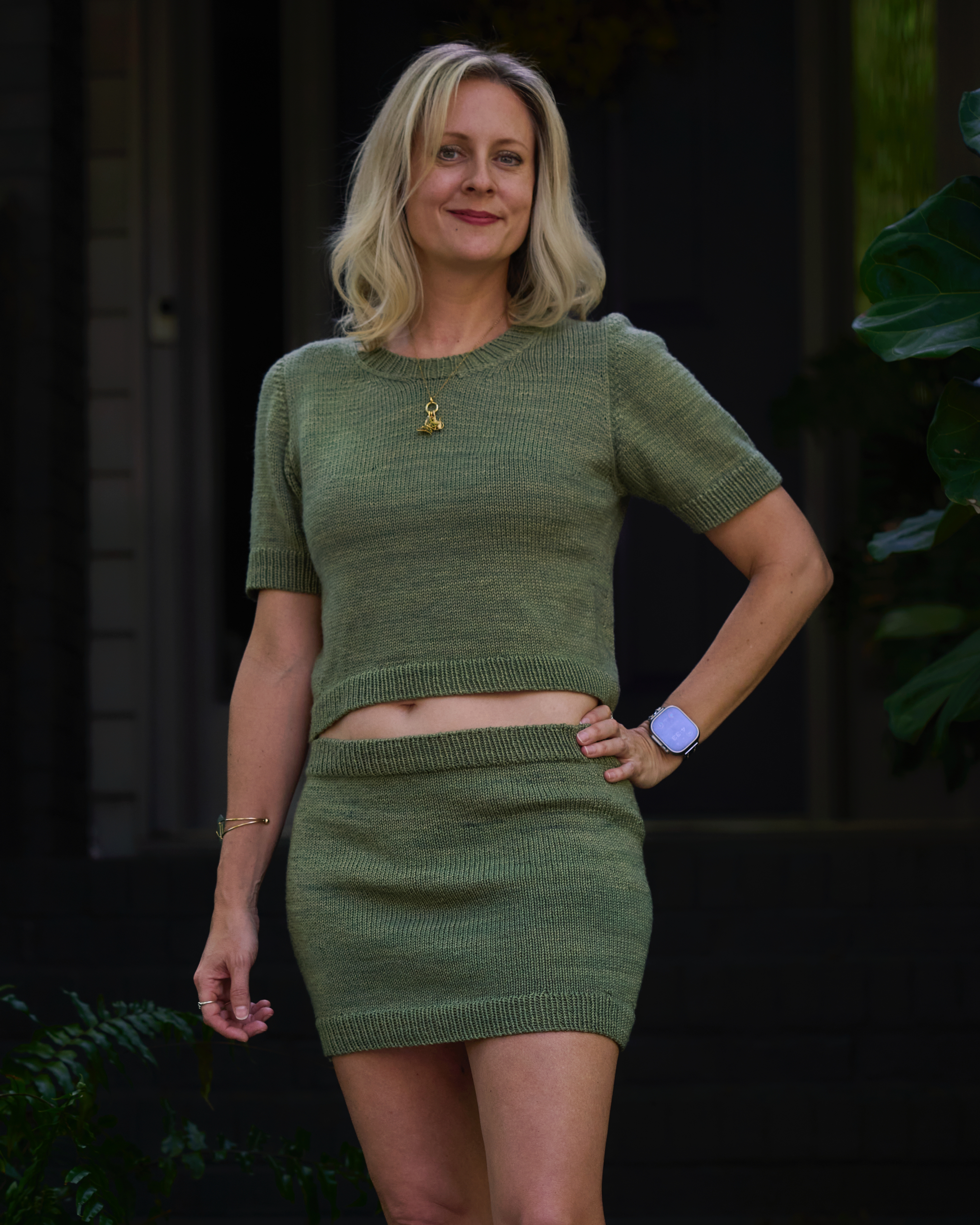 A woman wears a matching hand knit short-sleeved sweater and skirt set that is a warm sage green.